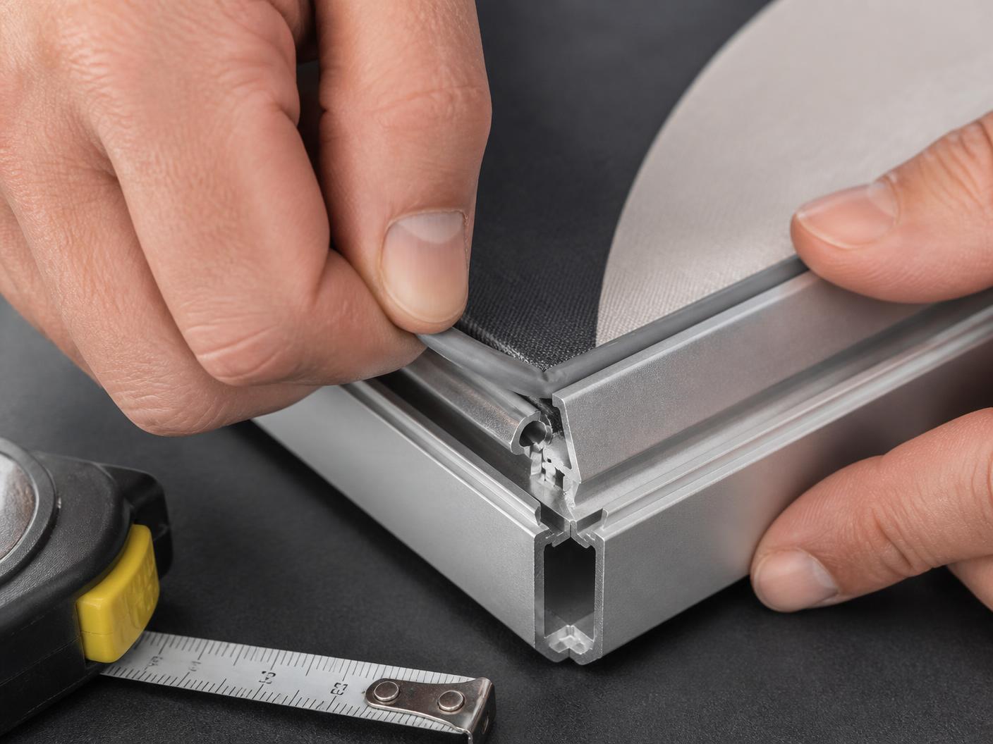 Close-up of a silicone edge strip being tucked into the channel of an aluminum SEG frame next to a measuring tape